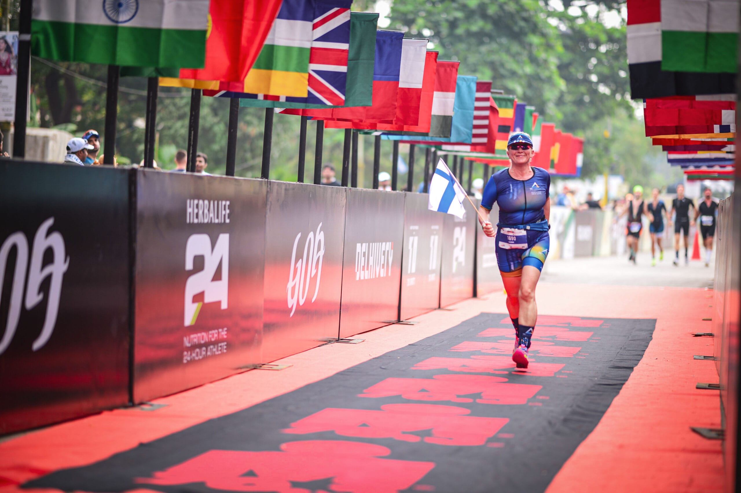 Nainen kilpa-asussaan juoksee kohti taustallaan eri maiden lippuja ja lehtipuita.